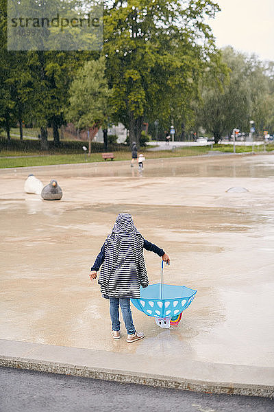 Rückansicht eines Mädchens im Regenmantel  das während der Regenzeit im Park mit einem Regenschirm spielt
