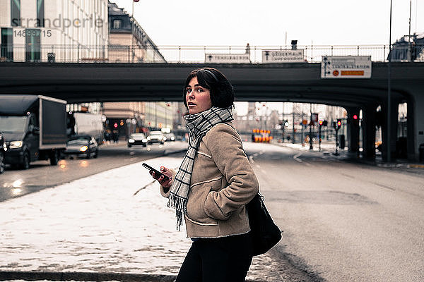 Seitenansicht einer jungen Frau  die im Winter beim Überqueren einer Straße in der Stadt ein Mobiltelefon in der Hand hält