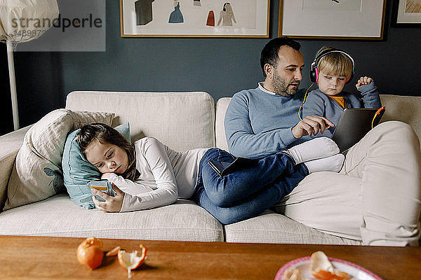 Mädchen benutzt Mobiltelefon  während der Vater der Schwester auf der Couch im Wohnzimmer zu Hause ein digitales Tablet zeigt