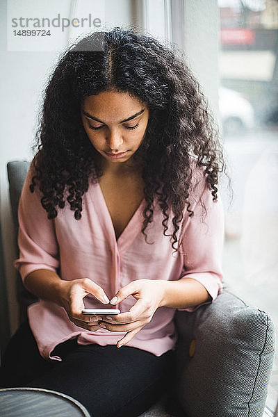 Junge Geschäftsfrau  die im Kreativbüro am Fenster sitzt und Textnachrichten von einem Smartphone aus verschickt