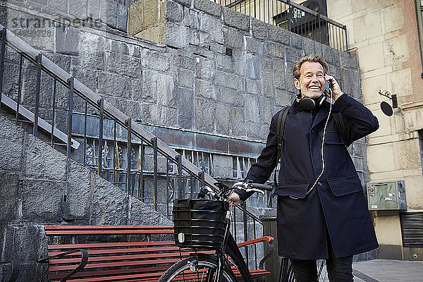 Lächelnder Mann spricht auf Smartphone mit Elektrofahrrad gegen Gebäude