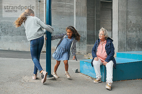 Lächelnde Großmutter betrachtet verspielte Enkelinnen  die sich auf dem Spielplatz um eine Stange drehen