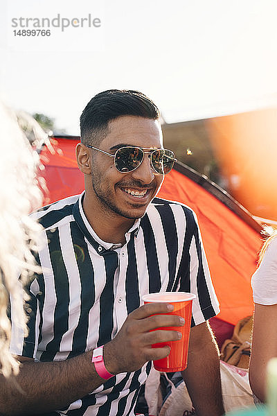 Lächelnder junger Mann mit Sonnenbrille trinkt während eines Musikfestivals an einem sonnigen Tag