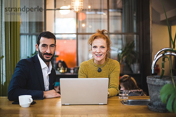 Porträt von lächelnden weiblichen und männlichen Geschäftskollegen  die mit Laptop am Tisch im Büro sitzen