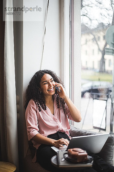 Lächelnde Jungunternehmerin spricht durch ein Smartphone  das im Büro auf dem Fensterbrett sitzt