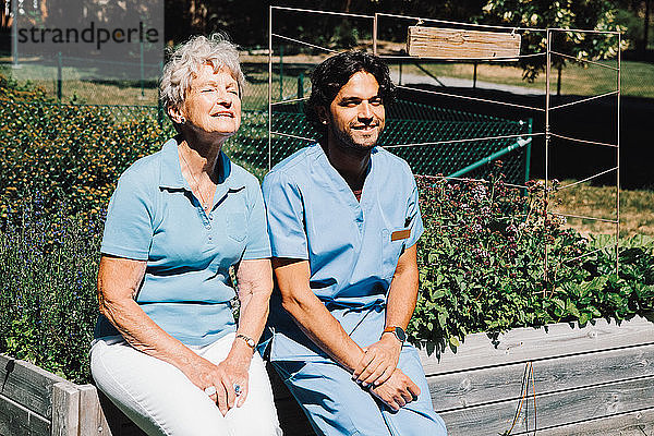 Lächelnde ältere Frau sitzt mit einem Pfleger auf einem Pflanzgefäß im Garten