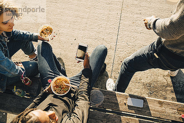 Schrägaufnahme eines Teenagers  der ein Smartphone benutzt  während er mit Freunden auf der Straße in der Stadt isst