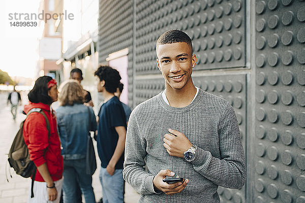 Porträt eines lächelnden Teenagers mit Handy in der Hand  während Freunde im Hintergrund auf der Straße stehen