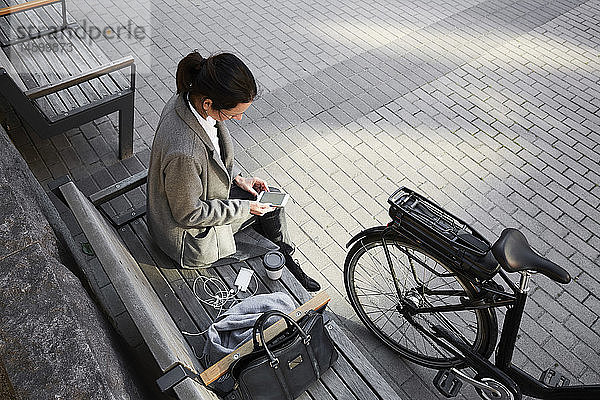 Schrägaufnahme einer Frau  die mit einem Smartphone im Internet surft  während sie in der Stadt auf einer Bank sitzt