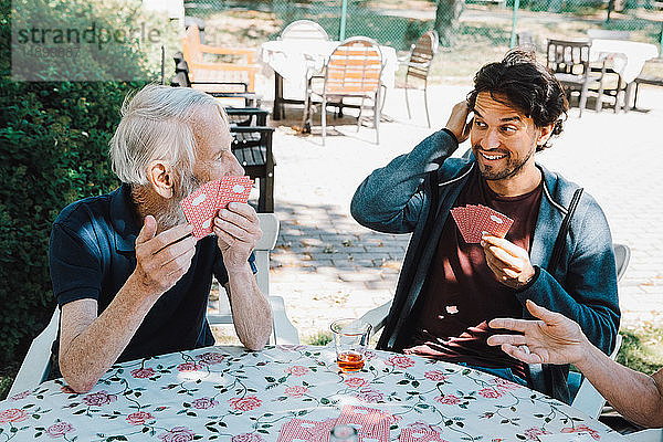 Lächelnder männlicher Hausmeister spielt Karten mit einem älteren Mann und einer älteren Frau am Tisch im Hinterhof