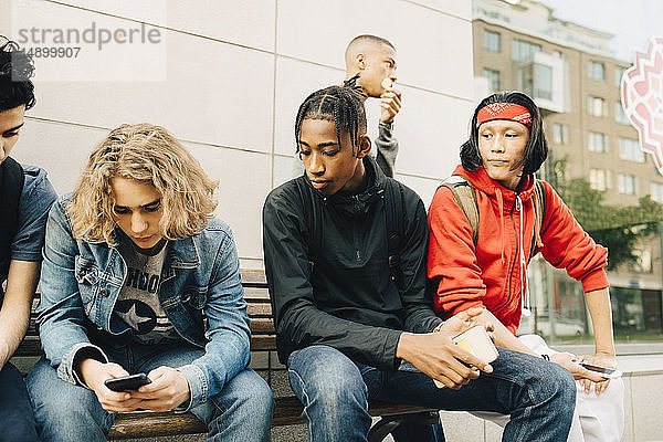 Junger Mann benutzt Mobiltelefon  während er bei Freunden auf der Bank am Bürgersteig sitzt
