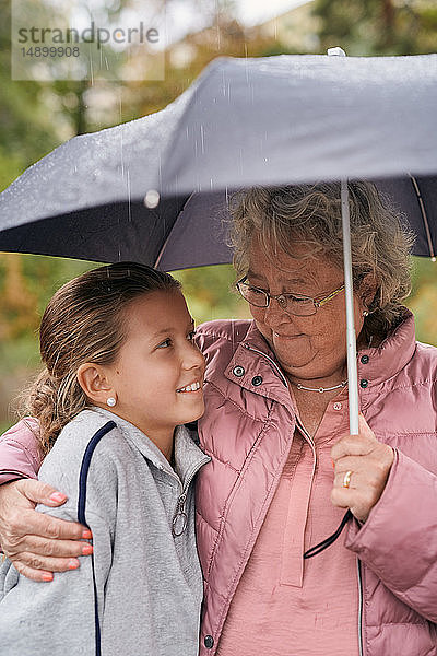 Großmutter umarmt Enkelin  während sie mit ihr im Park während der Regenzeit den Schirm teilt