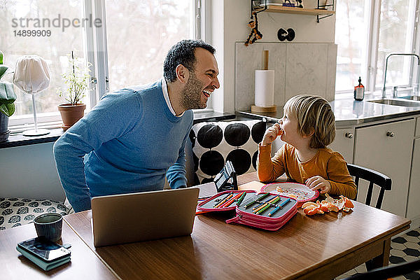 Verspielter Vater sieht Tochter an  während er zu Hause einen Laptop benutzt