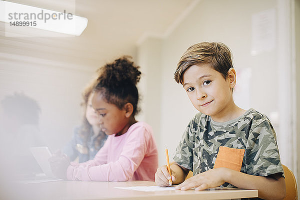 Porträt eines Jungen  der mit einem Freund am Schreibtisch im Klassenzimmer sitzt und auf Papier schreibt