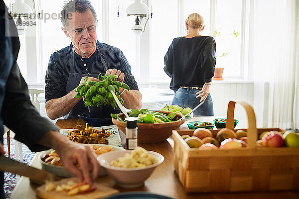 Reife Männer bereiten das Essen vor  während die Freundin zu Hause im Hintergrund steht