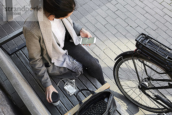 Schrägaufnahme einer Frau  die in der Stadt auf einer Bank sitzend per Mobiltelefon im Netz surft