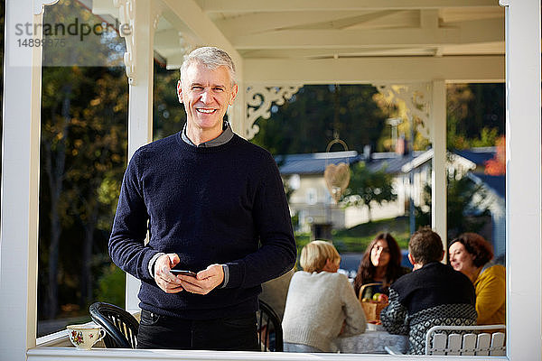 Porträt eines selbstbewusst lächelnden reifen Mannes  der auf der Veranda mit Freunden im Hintergrund steht