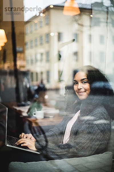 Porträt einer lächelnden jungen Berufstätigen  die mit einem Laptop durch ein Fenster im Büro sitzt