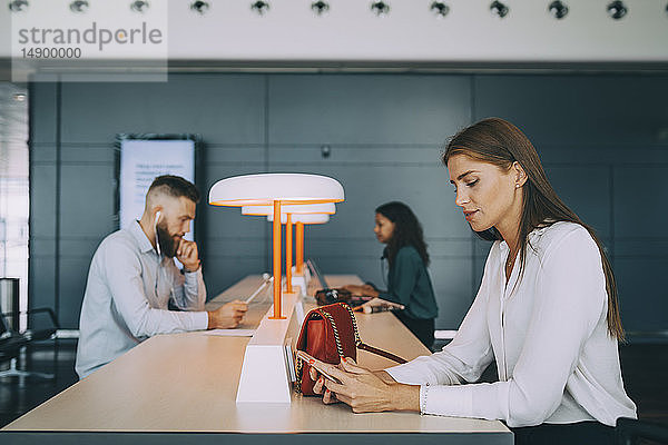 Junge Geschäftsfrau  die ein Smartphone benutzt  während sie mit multiethnischen Kollegen am Tisch sitzt und beim Abflug am Flughafen wartet a