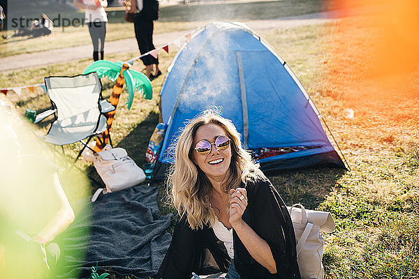 Hochwinkelansicht einer fröhlichen Frau mit Sonnenbrille  die im Sommer vor dem Zelt auf dem Feld sitzt