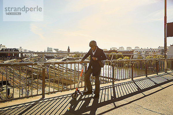 In voller Länge stehender junger Mann mit elektrischem Schieberoller an einem Geländer auf einer Brücke in der Stadt
