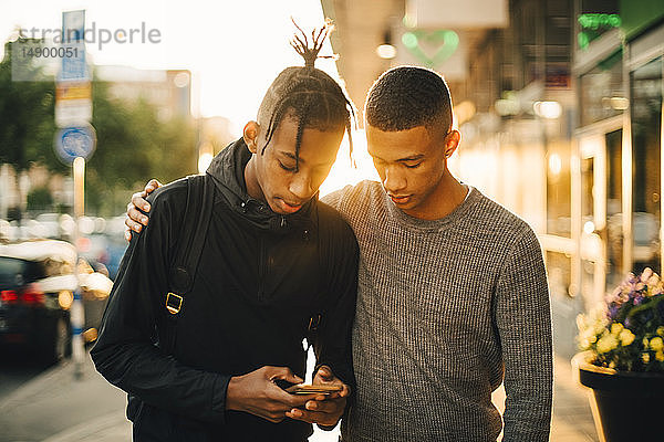 Teenager  der ein Mobiltelefon benutzt  während er einem Freund bei Sonnenuntergang auf der Straße in der Stadt zur Seite steht