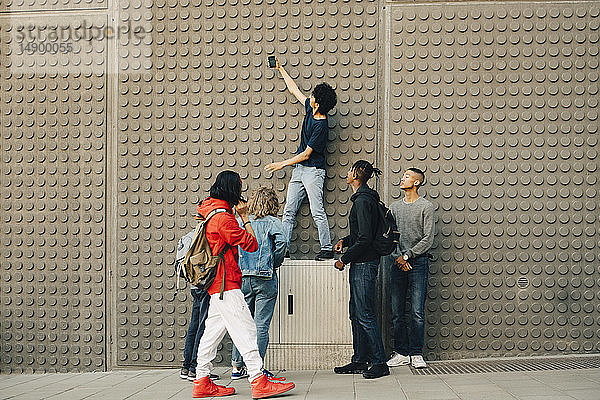 Männliche Freunde in voller Länge beim Selfie mit dem Handy  während sie in der Stadt an der Mauer stehen