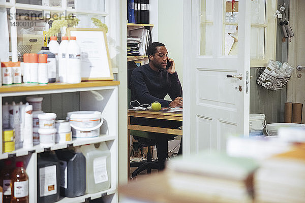 Selbstbewusster junger männlicher Besitzer  der am Schreibtisch im Möbelgeschäft sitzt und mit einem Smartphone telefoniert
