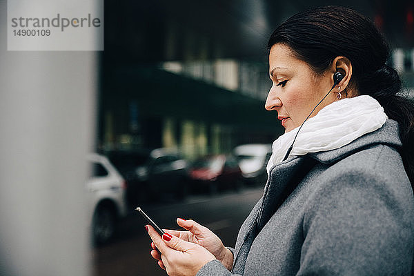 Seitenansicht einer Geschäftsfrau  die mit einem Mobiltelefon Musik hört  während sie in der Stadt auf der Straße steht