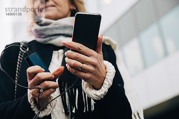 Geschäftsfrau hält ein Mobiltelefon mit Ladegerät in der Hand  während sie sich gegen ein Gebäude in der Stadt stellt