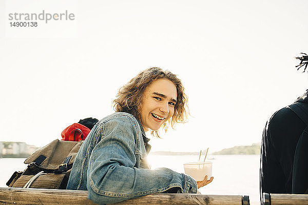 Porträt eines fröhlichen jungen Mannes  der eine Mahlzeit in einem Behälter hält  während er mit einem Freund an einem sonnigen Tag auf der Promenade sitzt