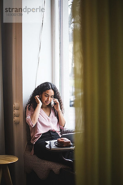 Junge Berufstätige  die im Büro auf der Fensterbank sitzt und per Smartphone telefoniert