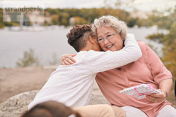 Enkel umarmt lächelnde Großmutter bei der Entgegennahme eines Geschenks am Seeufer im Park während eines Picknicks