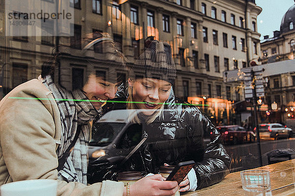 Lächelnde Freundinnen  die ein Mobiltelefon benutzen  während sie in einem Café sitzen und durch ein Fenster gesehen werden