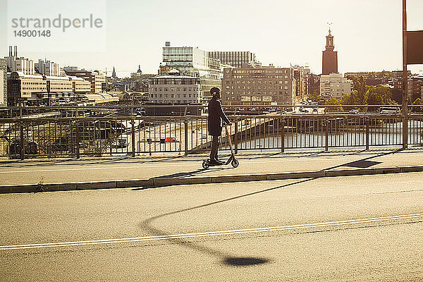 Ganzflächige Seitenansicht eines jungen Mannes auf einem Elektroschubroller auf einer Brücke in der Stadt