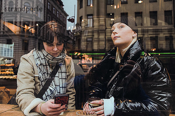 Nachdenkliche junge Frau  die durch ein Glasfenster schaut  während ein Freund im Winter im Café ein Mobiltelefon benutzt