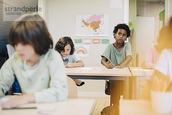 Neugieriger Junge malt mit Freunden am Schreibtisch im Klassenzimmer