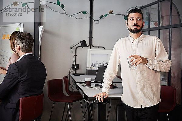 Nachdenklicher männlicher Unternehmer hält ein Trinkglas in der Hand  während er im kreativen Büro am Schreibtisch steht