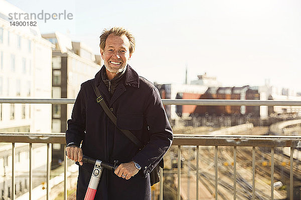 Porträt eines lächelnden reifen Mannes  der mit einem elektrischen Schubsroller auf einer Brücke gegen den Himmel in der Stadt steht