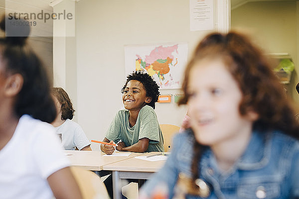 Lächelnder Junge sitzt mit Freunden am Schreibtisch im Klassenzimmer