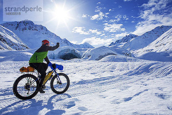 Frau auf dem Fatbike vor dem Skookum-Gletscher  Chugach National Forest  Alaska  an einem sonnigen Wintertag  die Faust pumpt  während sie vorbeifährt  Süd-Zentral-Alaska; Alaska  Vereinigte Staaten von Amerika