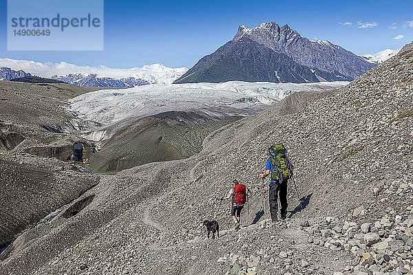 Ehepaar und ihr Hund wandern auf einer Gletschermoräne in Richtung Root Glacier  mit Donoho Peak und den Wrangell Mountains im Hintergrund  Wrangell-St. Elias National Park  Süd-Zentral-Alaska; Kennicott  Alaska  Vereinigte Staaten von Amerika
