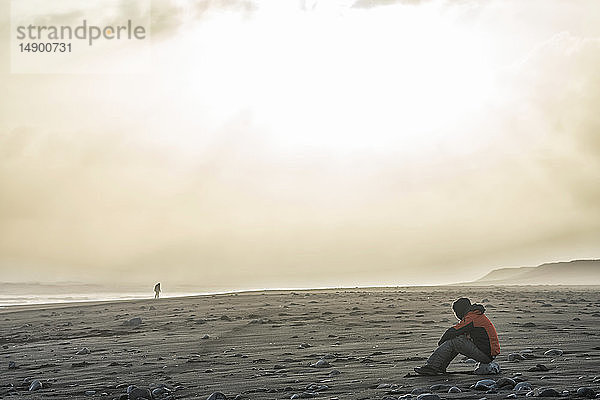Eine Person sitzt am Ufer von Südisland und beobachtet eine andere Person  die in der Ferne spazieren geht. Stimmungsvolle Beleuchtung erhellt die Landschaft; Island