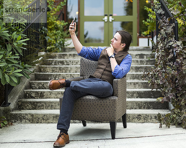Ein junger Mann sitzt in einem Sessel im Freien und benutzt sein Smartphone  um ein Selbstporträt aufzunehmen; Bothell  Washington  Vereinigte Staaten von Amerika
