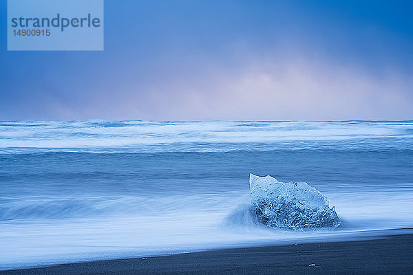 Eisstück  das von den Wellen entlang der Südküste Islands getroffen wird  mit stürmischem Himmel dahinter; Island