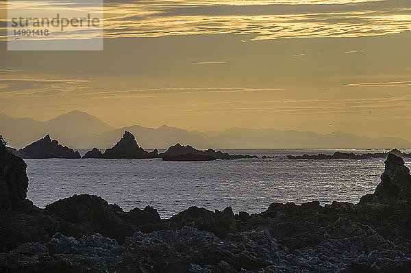 Glühende  goldene Wolken und Silhouetten von Felsformationen und der zerklüfteten Küstenlinie entlang der Südküste bei Sonnenuntergang; Wellington  Neuseeland