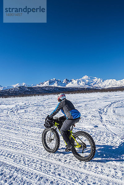 Ein Mann fährt an einem sonnigen Wintertag mit einem Fatbike auf dem Chulitna Bluff Trail. Die Alaska Range und der 20.230' hohe Mount Denali (McKinley) sind im Hintergrund zu sehen  Süd-Zentral-Alaska; Alaska  Vereinigte Staaten von Amerika