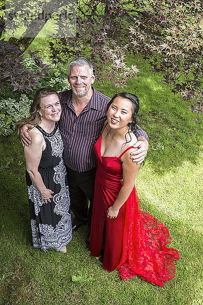 Porträt einer High-School-Absolventin in einem roten Gesellschaftskleid  die mit ihren Eltern in einem Hof steht; Chilliwack  British Columbia  Kanada
