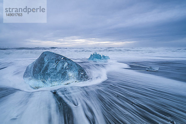 Ein großer Eisblock liegt an der Küste von Südisland  während Wellen an die Küste schlagen; Island