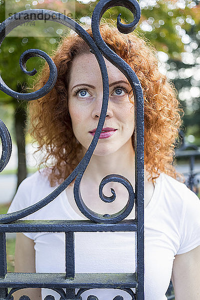 Porträt einer Frau mit rotem  lockigem Haar und strahlend blauen Augen  die hinter einem schmiedeeisernen Tor steht und nach oben schaut; Burnaby  British Columbia  Kanada